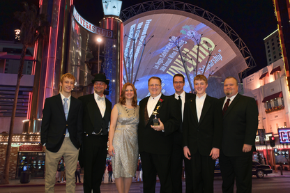 Seven men in formal evening attire pose together on a city street at night in front of a brightly lit Las Vegas club and arched building structure. Five wear black suits, one wears a black tuxedo with a white shirt and bow tie and a black hat, one wears a silver patterned dress, and one in the center holds a trophy.