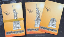 Three orange notebooks with black and white illustrations of the Statue of Liberty and other landmarks on a dark surface.