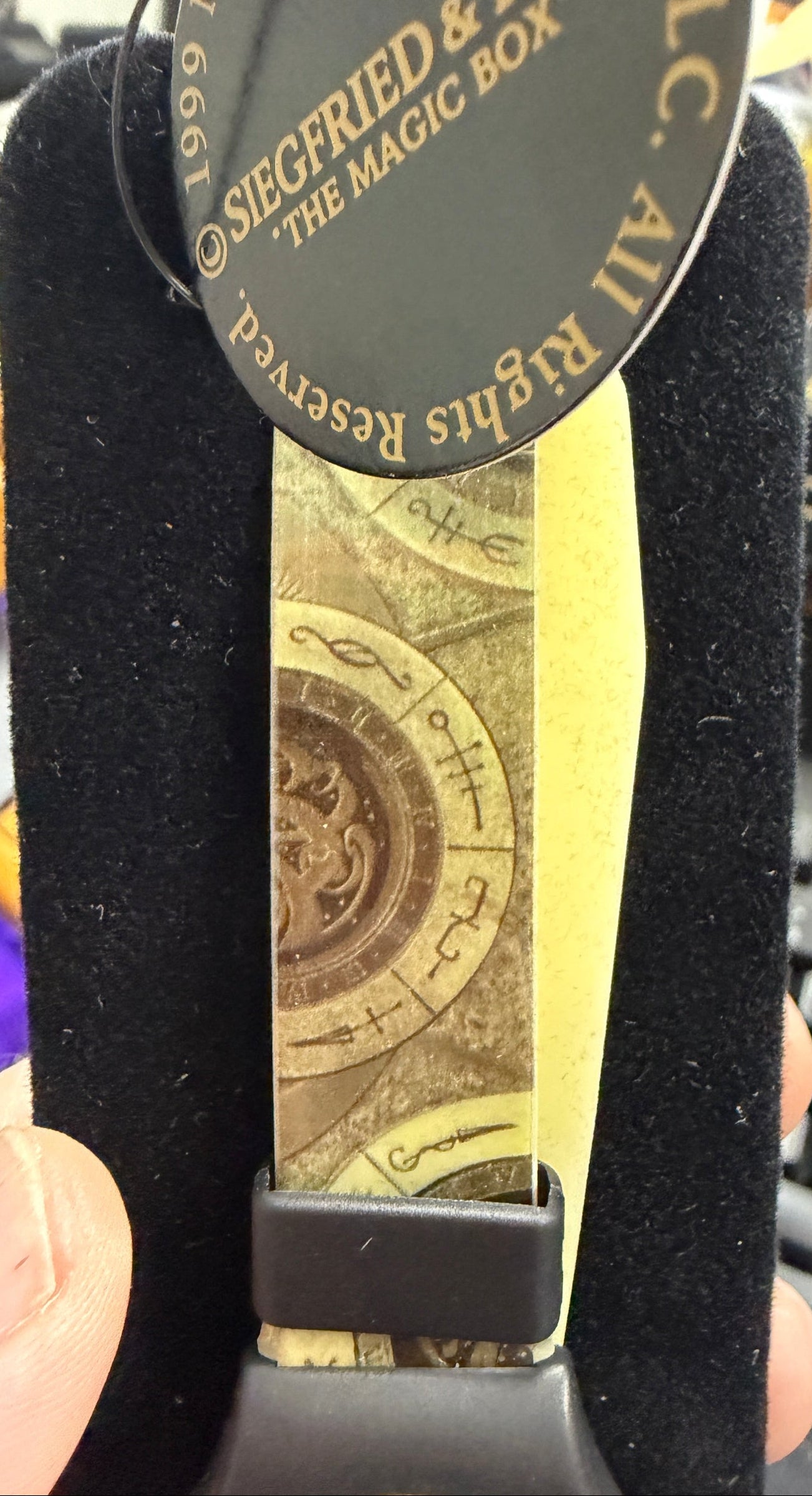 Black strap with a circular tag and printed design, held by a hand with a blurred background.