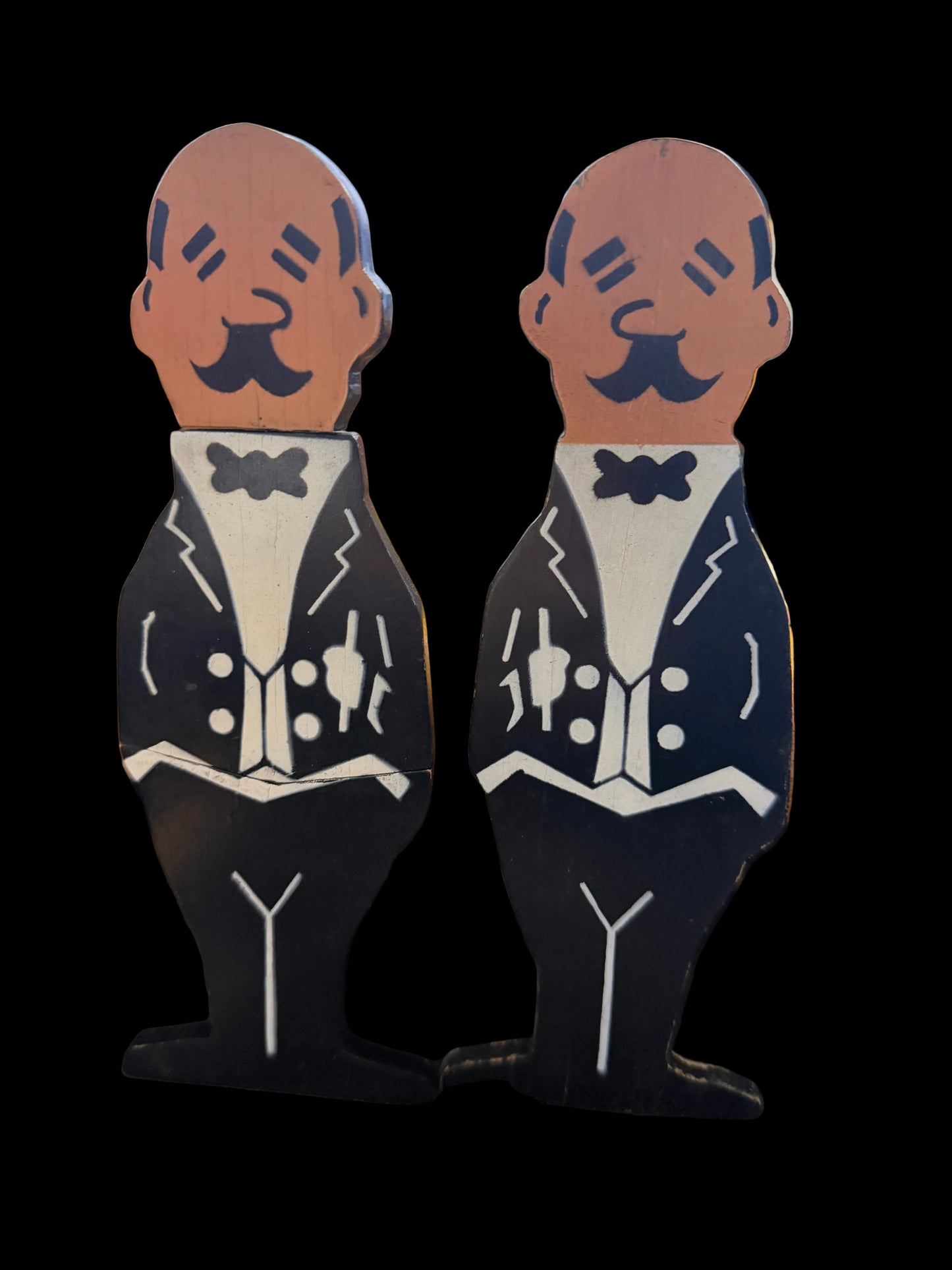 Wooden cutout figures of two men in suits attached to a wooden stick, with a blurred background.