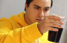Person in a yellow hoodie holding a glass of dark liquid with a logo in the corner.