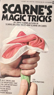Book cover of 'Scarne's Magic Tricks' with a hand holding a piece of paper over a card.