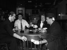 Black and white photograph of four men gathered around a round wooden table in a dimly lit bar or lounge, holding cards and smoking cigars.