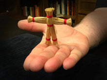 Hand holding a small straw figure with red bands against a blurred bookshelf background