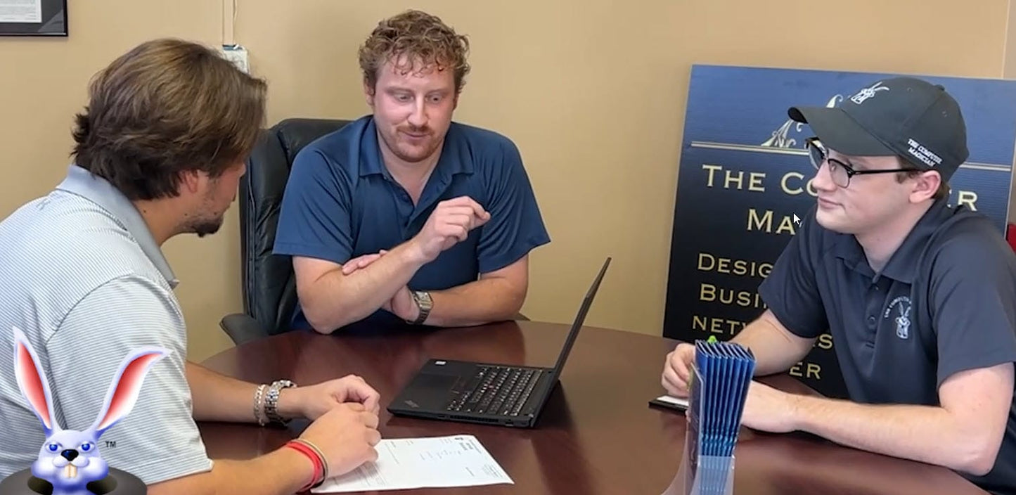 Three men sitting around a table with a laptop, discussing something.