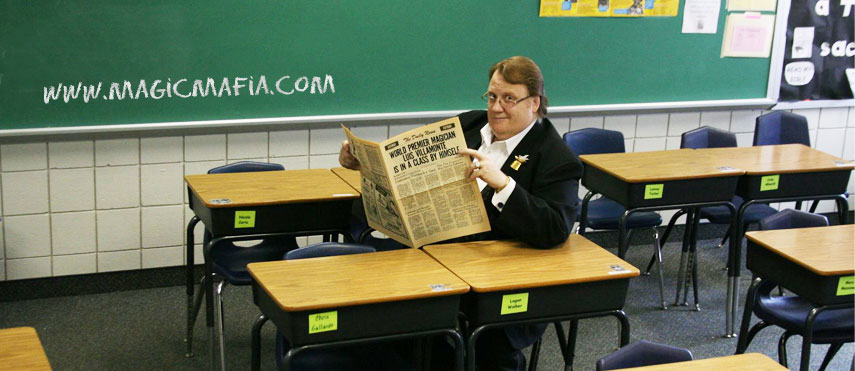 Person sitting at a desk in a classroom reading a newspaper, with a green chalkboard and desks in the background.