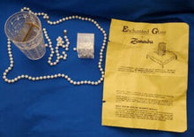 Glass cup with pearls, small box, and yellow 'Enchanted Glass' Zoraida packet on a blue background