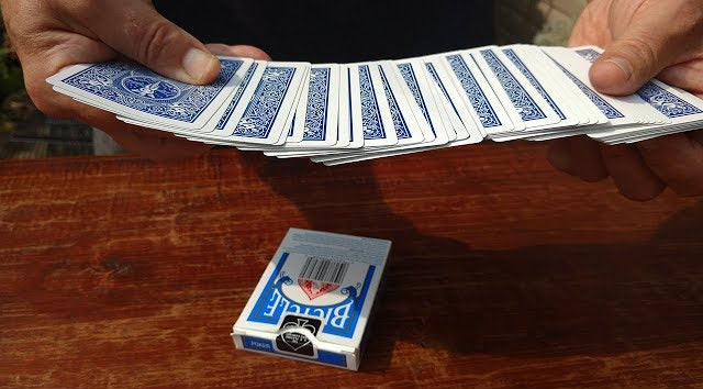 Person holding a deck of blue and white playing cards on a wooden surface with a deck of cards in the background.