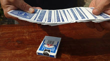 Person holding a deck of blue and white playing cards on a wooden surface with a deck of cards in the background.