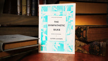 Book titled 'The Sympathetic Silks' by Lewis Ganson on a wooden surface with books in the background.